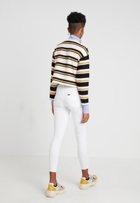 Suéter de canalé a rayas en negro, amarillo y blanco con diseño corto sobre una camisa de color lila; combinado con jeans ajustados blancos y zapatillas multicolor.