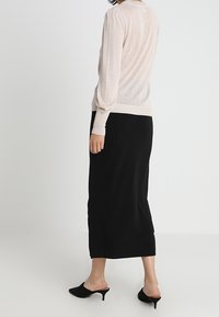 Kvinna i en genomskinlig beige långärmad topp, svart midikjol och svarta klackskor med kitten heel, står framför en vit bakgrund.