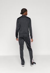 Μαύρο φόρμα με μπλούζα με φερμουάρ και παντελόνι που ταιριάζει. Διαθέτει ροζ ρίγες-λεπτομέρειες κατά μήκος των μανικιών και των πλευρών. Λείο, ελαφρύ ύφασμα.