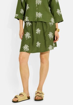 Femme portant une jupe et un haut verts avec des imprimés de palmiers blancs, des sandales beiges à boucle, ainsi qu'un bracelet et des bagues en or sur fond blanc.