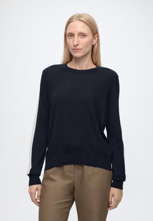 Blonde Frau mit einem schwarzen Langarmpullover mit weißen Streifen an den Ärmeln und brauner Hose, die vor einem schlichten hellen Hintergrund steht.