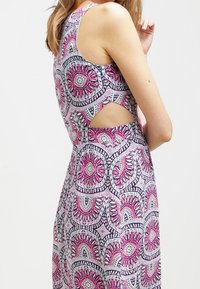 Kvinna i ärmlös klänning med rosa, vita och svarta cirkulära blommönster och med en sidoklippning ovanför midjan.