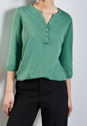 Personne portant un t-shirt henley vert à trois boutons avec des manches trois-quarts et un pantalon noir, debout devant un fond clair.