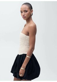 Top beige tanpa tali dengan tekstur menggumpal, dipadukan dengan rok hitam yang voluminous. Aksesoris perhiasan emas melengkapi penampilan.