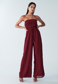 Bordo brez naramnic kombinezon z nabrano zgornjim delom, ježkom v pasu in širokimi hlačnicami; iz lahkotnega, tekočega blaga.