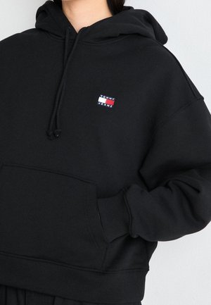 Musta huppari etutaskulla ja pienellä Tommy Jeans -logolla vasemmassa rinnassa, henkilön päällä, jolla käsi taskussa.