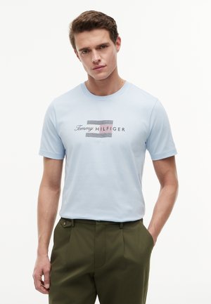 Ung mand iført en lyseblå Tommy Hilfiger t-shirt med logo og olivengrønne bukser, stående med den ene hånd i lommen mod hvid baggrund.
