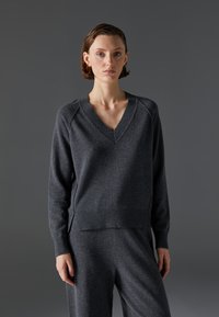 Temno siva pulover s V-izrezom iz mehkega pletenega materiala, z raglan rokavi in ribanim robom, v kombinaciji z ujemajočimi sivimi hlačami.