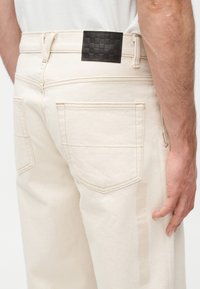 Κοντινό πλάνο ενός ατόμου που φοράει τζιν παντελόνια μπεζ με πίσω τσέπ ες και μια μαύρη δερμάτινη ταμπέλα με καρό σχέδιο στη ζώνη.