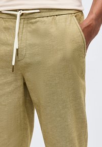 Primo piano di una persona che indossa pantaloni chiari verde oliva con coulisse e mano in tasca, abbinati a una maglia a costine di colore chiaro.