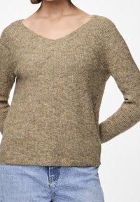 Pull en laine beige avec col en V, motif en maille texturée et manches longues, associé à un jean bleu clair. Design simple et décontracté.