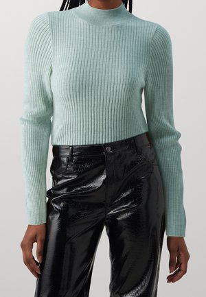 Kobieta nosząca jasnoniebieski prążkowany sweter z długim rękawem oraz czarne, błyszczące spodnie z wysokim stanem wykonane ze sztucznej skóry z wzorem krokodyla.