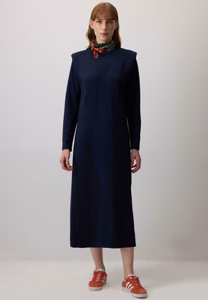 Robe en jersey bleu marine à manches longues avec un col haut et des accents sur les épaules, associée à des baskets orange et une écharpe à motifs autour du cou.