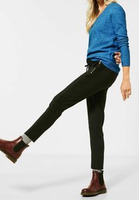 Kvinna med långt blont hår som bär en blå tröja, svarta byxor med dragkedjefickor, gråa strumpor och bruna läderankelstövlar, lyfter ett ben.