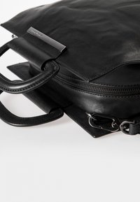 Musta nahkalaukku, jossa on sileä pinta, vetoketjusuljenta ja kaksi pyöreää kahvaa. Laukussa on metallikoriste ja irrotettava hihna tasaisella pohjalla.