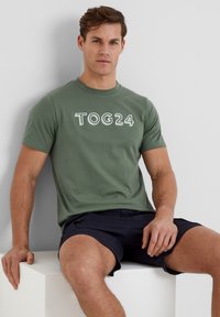 Olive green short-sleeve t-shirt with white "TOG24" graphic print. Soft fabric, classic crew neck, worn with navy shorts, sitting on a white surface.
