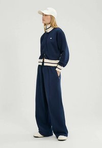 Marineblauer Reißverschluss-Pullover mit cremefarbenen Streifen an Bündchen und Saum, kombiniert mit locker sitzenden marineblauen Jogginghosen. Weißes Cap. Weicher, strukturierter Stoff.