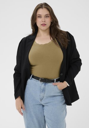 Femme portant un blazer noir sur un débardeur vert olive rentré dans un jean taille haute bleu clair avec une ceinture noire, posant avec une main dans la poche.