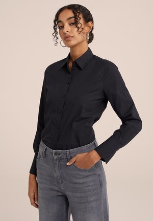 Chemise noire à boutons avec une coupe ajustée, manches longues et col droit, associée à un pantalon en denim gris clair. Texture de tissu lisse.