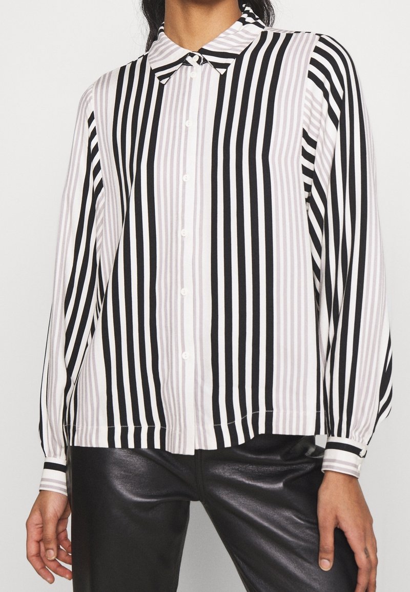 Femme portant une chemise noire et blanche à rayures verticales avec boutons, manches longues et un pantalon en cuir noir.