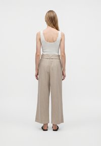 Mujer de pie de espaldas, con una camiseta sin mangas blanca y pantalones beige de cintura alta y pernera ancha, con sandalias negras sobre un fondo liso.
