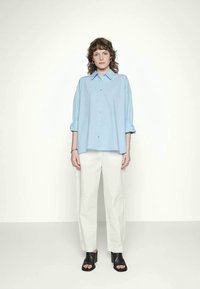 Camisa azul claro de botões com mangas arregaçadas, combinada com calças brancas e sapatos de salto abertos pretos. Design simples e de corte relaxado.