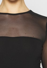 Svart topp med en genomskinlig mesh-övre del och en solid svart nedre del. Halsringningen är rundad med kontrasterande tyger och texturer.