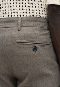 Gros plan sur la taille d'une personne portant un pantalon marron texturé avec une poche arrière boutonnée et un pull en maille crème.