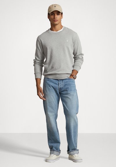 Sivi dukserica s okruglim izrezom, svijetloplave traperice i bež kapa s logom. Bijele tenisice upotpunjuju odjevnu kombinaciju.