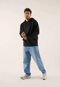 Schwarzer Kapuzenpullover mit langen Ärmeln, kombiniert mit hellblauen lockeren Jeans und grau-weißen Turnschuhen. Einfaches Design, Material aus Baumwollmischung.