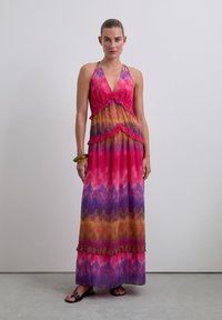 Vrouw staat in een mouwloze halterhals maxi-jurk met een roze, paarse en oranje tie-dye patroon en ruches, zwarte sandalen en een groene armband.