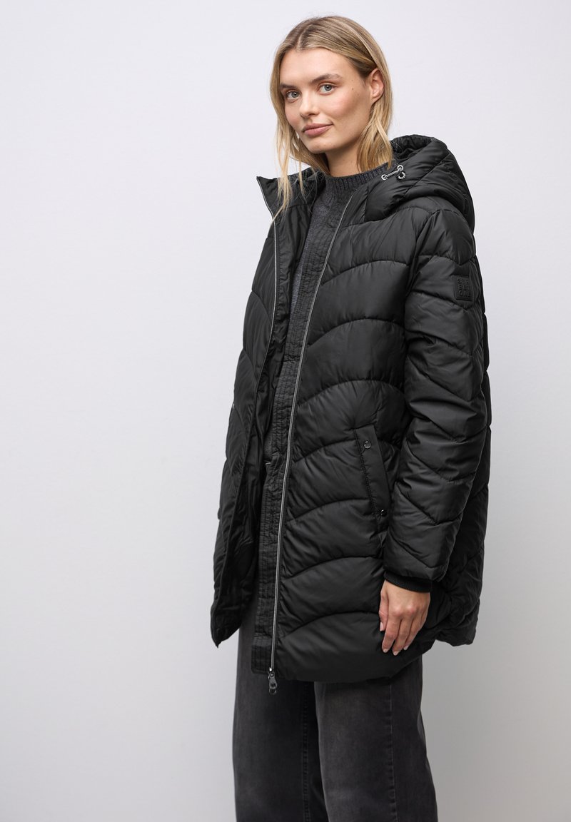 Giacca puffa nera con zip frontale, cappuccio e tasche laterali. Caratterizzata da un design trapuntato e una texture morbida, arriva fino a metà coscia.