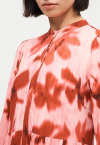 Γυναίκα που φοράει ροζ φόρεμα με κόκκινα λουλουδάτα σχέδια και ψηλό γιακά, αποκαλύπτοντας το επάνω μέρος του στήθους και τον λαιμό.