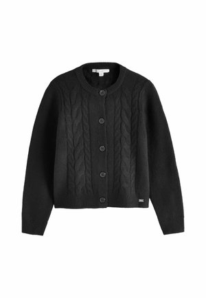 Nahaufnahme eines schwarzen Strickpullovers mit Metallplakette, getragen mit dunkelgrauer Denim-Jeans und einer teilweise sichtbaren Hand an der Seite.