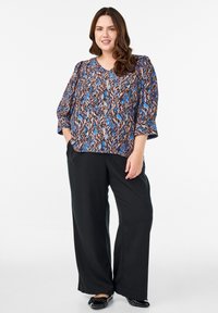 Mønstret bluse i flere farver med V-hals og trekvartærmer, parret med brede sorte bukser og sorte flade sko. Blusen har et svungent mønster.