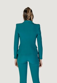 Teal oblek s jedním rozventilovaným zadním dílem, strukturovanými rameny a dlouhými rukávy, doplněný o odpovídající fitted kalhoty. Hladká textura látky.