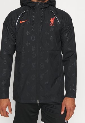 Czarna kurtka z kapturem z subtelnymi ciemnymi wzorami, czerwone logo Liverpool FC na piersi, czerwony znak Nike, pełny zamek błyskawiczny, noszona przez osobę z widocznymi rękami.