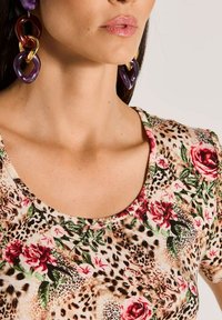 Frau mit einem floralem und Leopardenmuster-Oberteil, die große Ohrringe aus lila, braunen und goldenen Kettengliedern trägt.