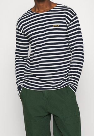 Hombre con camiseta de manga larga a rayas horizontales en azul marino y blanco con el texto "amour" en el pecho y pantalones verde oscuro con las manos en los bolsillos.