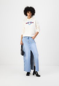 Light cream hoodie with "NEW YORK" graphic, paired with a mid-length denim skirt featuring a front slit. Black ankle boots and a small bag.