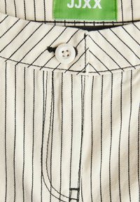 Tessuto bianco con righe verticali nere, caratterizzato da una chiusura frontale con bottoni, un'etichetta verde del marchio e cuciture dettagliate.