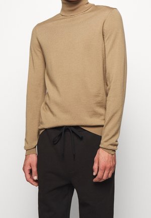Homme portant un pull à col roulé beige rentré dans un pantalon noir à cordon, sur fond blanc.