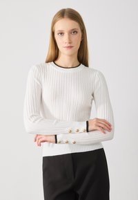 Pull blanc côtelé à manches longues, avec des bordures noires au niveau du cou et des poignets, et des accents de boutons dorés sur les bords des manches.