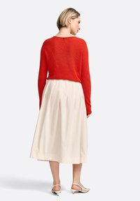 Pull en tricot rouge à manches longues, associé à une jupe plissée couleur crème. Comprend un petit bouton noir à l'arrière du pull.