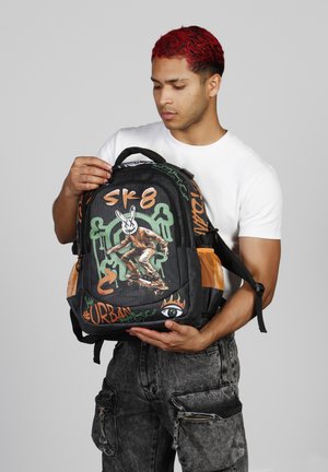 Mochila negra con arte estilo graffiti, bolsillos laterales de malla naranja y detalles verdes. Presenta un diseño de patinador y elementos de texto urbano.