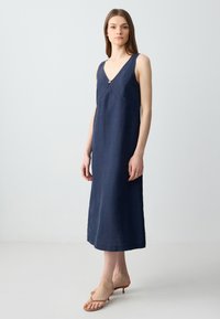 Marineblaues Leinenkleid mit V-Ausschnitt, ärmellosem Design und einem dekorativen Metallelement an der Vorderseite. Bodenlang mit einer lockeren Passform.