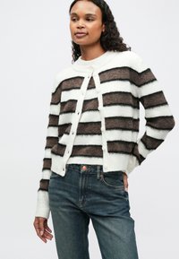 Cardigan a righe nei toni crema, marrone e nero, con chiusura a bottoni, colletto a costine e polsini. Indossato su jeans scuri.