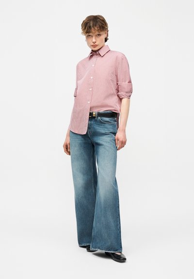 Άτομο που φοράει ροζ πουκάμισο με μανίκια γυρισμένα, φαρδιά ξεβαμμένα μπλε τζιν με μαύρη ζώνη και μαύρα επίπεδα παπούτσια, όρθιο μπροστά σε λευκό φόντο.