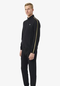 Jonge man in een zwarte trainingspak met een volledige rits en gouden strepen langs de mouwen, staande tegen een eenvoudige witte achtergrond.