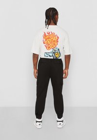 Witte T-shirt met korte mouwen, voorzien van een kleurrijke vlam- en aanstekerafbeelding op de rug, gecombineerd met zwarte cargobroek en witte sneakers.